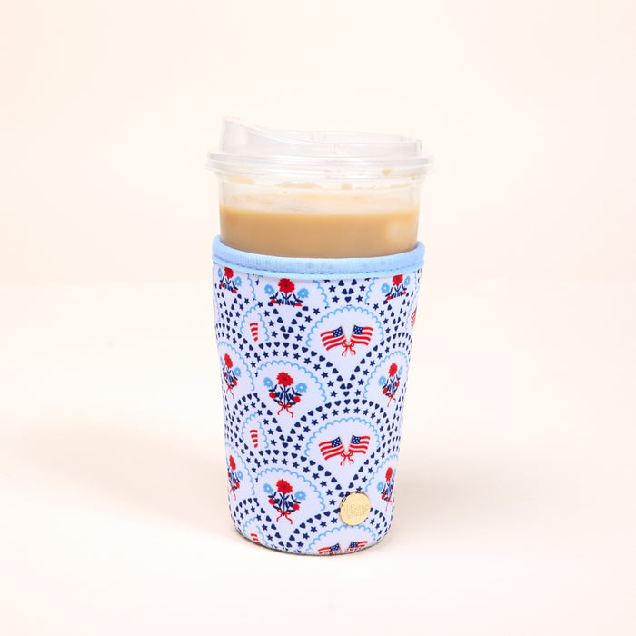 A clear plastic cup of iced coffee sits in The Darling Effect's Large-All-American Darling Sip Savvy Drink Sleeve, featuring flags, hearts, and flowers on light blue, against a plain light background.