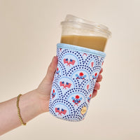 A hand holds an iced coffee in a plastic cup wrapped in The Darling Effect’s Large-All-American Darling Sip Savvy Drink Sleeve, featuring American flags and red, white, and blue patterns. A gold beaded bracelet is on the wrist.