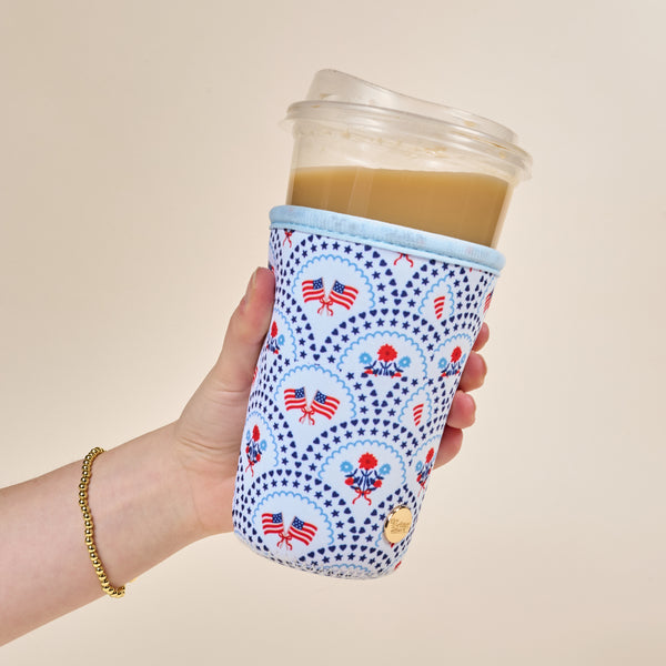 A hand holds an iced coffee in a plastic cup wrapped in The Darling Effect’s Large-All-American Darling Sip Savvy Drink Sleeve, featuring American flags and red, white, and blue patterns. A gold beaded bracelet is on the wrist.