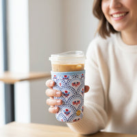A smiling woman in a white sweater holds an iced coffee with The Darling Effect’s Large-All-American Darling Sip Savvy Drink Sleeve, sitting at a wooden table in a bright room.