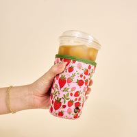 A hand holds an iced drink in a clear cup with The Darling Effect's Large-Strawberry Fields Sip Savvy Drink Sleeve, featuring a pink strawberry pattern on soft neoprene. A gold bracelet is worn; background is light beige.