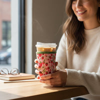 A woman in a white sweater sits by a window, smiling and holding a coffee cup with The Darling Effect’s Large-Strawberry Fields Sip Savvy Drink Sleeve. Glasses and notebooks rest on the table beside her.