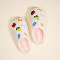 The Darling Effect’s Italian Summer Fuzzy Slippers, shown on a light beige background, are white with a soft pink insole. Embroidered lemons, candy, bows, and strawberries add playful charm along with a visible label.