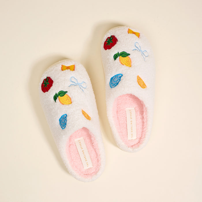 The Darling Effect’s Italian Summer Fuzzy Slippers, shown on a light beige background, are white with a soft pink insole. Embroidered lemons, candy, bows, and strawberries add playful charm along with a visible label.