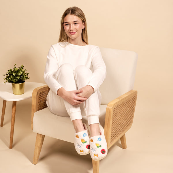 A young woman in white loungewear sits cross-legged on a beige armchair, smiling softly. She wears The Darling Effect’s Italian Summer Fuzzy Slippers with colorful fruit designs. A small potted plant rests on the side table beside her.