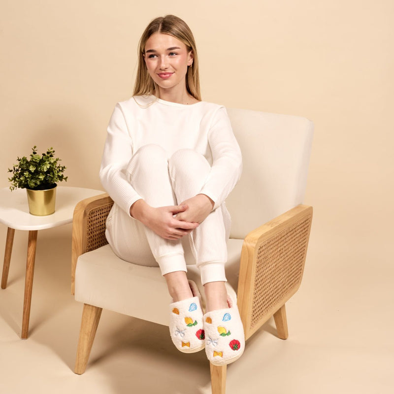 A young woman in white loungewear sits cross-legged on a beige armchair, smiling softly. She wears The Darling Effect’s Italian Summer Fuzzy Slippers with colorful fruit designs. A small potted plant rests on the side table beside her.