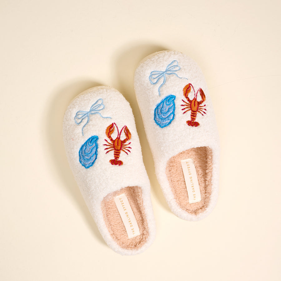 Seaside Haven Fuzzy Slippers by The Darling Effect feature white plush material, blue oyster and orange lobster embroidery, light blue bows, a pink sherpa lining—cozy slippers perfect for lounging on a cream background.