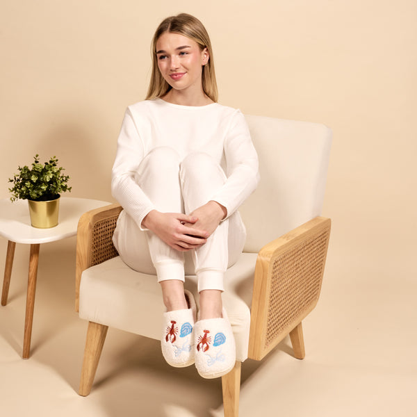 A woman in white loungewear, smiling and cross-legged in a beige armchair, wears The Darling Effect's Seaside Haven Fuzzy Slippers with blue and red embroidery. Beside her is a small plant on a white table against a beige background.