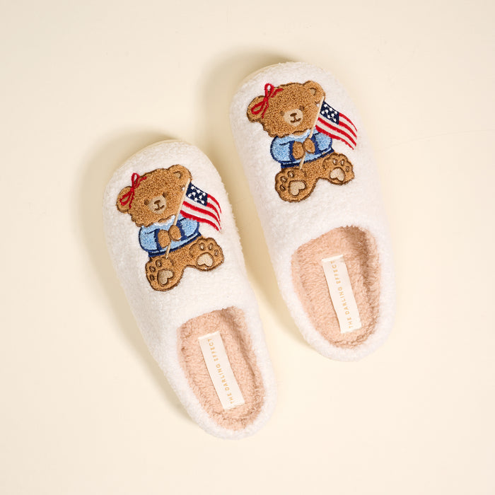 The Darling Effect’s Star Spangled Bear Fuzzy Slippers are white, plush, and feature embroidered teddy bears with American flags plus paw prints on a light cream background.