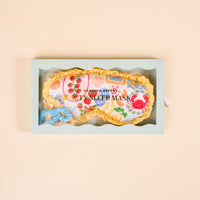 The Darling Effect Ciao Bella Beauty Sleep Mask set features a floral satin mask with yellow trim, blue satin scrunchie, and comes in a light green box with clear window. "Beauty Sleep Mask" is printed on the mask.