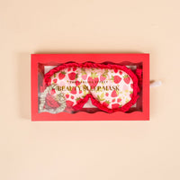 The Darling Effect's Strawberry Fields Beauty Sleep Mask features white satin with red ruffles and a strawberry print, packaged in a red box with a clear lid. This set also includes a matching scrunchie for extra comfort.