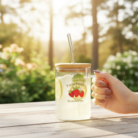 A hand holds The Darling Effect’s Farm Fresh Strawberries Square Glass Cup, filled with lemonade, lemon slices, mint, ice, a bamboo lid, and a glass straw. The cup rests on a wooden table outdoors surrounded by greenery and sunlight.