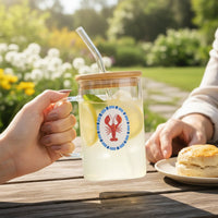 A hand holds The Darling Effect’s Lobster Square Glass Cup, filled with lemonade, lemon slices, mint, a bamboo lid, and a glass straw. A red lobster design decorates the cup. Another hand and a biscuit plate sit on an outdoor garden table.