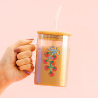 A hand holds The Darling Effect Off the Vine Square Glass Cup, featuring a tomato vine design and bamboo lid, filled with a caramel drink and topped with a glass straw.