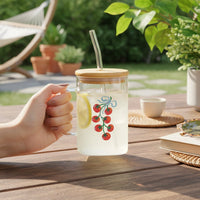 A hand holds The Darling Effect's Off the Vine Square Glass Cup, featuring a cherry tomato design, iridescent glass, bamboo lid, and glass straw. The mug with lemonade and ice shines on an outdoor wooden table.