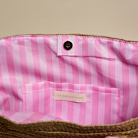 A close-up of the Dolce Vita Straw & Sun Tote by The Darling Effect shows its pink and white striped lining, snap closure, and an interior pocket branded with The Darling Effect.