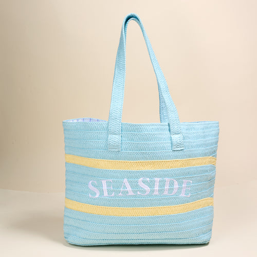 The Darling Effect Seaside Straw & Sun Tote features light blue and yellow stripes with "SEASIDE" embroidered in white—a lightweight, stylish choice for your next vacation.
