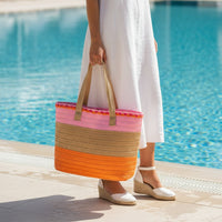 A woman in a white dress and white wedge sandals stands by a pool, holding The Darling Effect’s Tropical Sunset Straw & Sun Tote—a large striped bag featuring shades of orange, brown, pink, and tan.