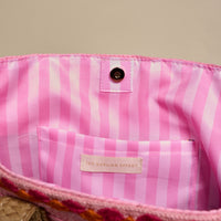 The image shows the inside of The Darling Effect's Tropical Sunset Straw & Sun Tote, lined with pink and white stripes. It features a small pocket with the brand label and a snap button closure—perfect for your next vacation.