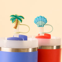 The Island Escape Straw Topper Set by the-darling-effect includes two leak-resistant, colorful tumblers—one blue with a palm tree topper and one red with a blue seashell topper—perfect for fun drinkware against a light beige background.