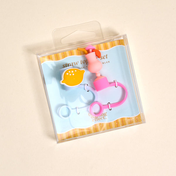The Darling Effect’s Limoncello Love Straw Topper Set includes leak-resistant pink and yellow lemon-shaped and pink ring silicone toppers, adding fun to your drinkware accessories. Displayed on a light blue and yellow striped background.