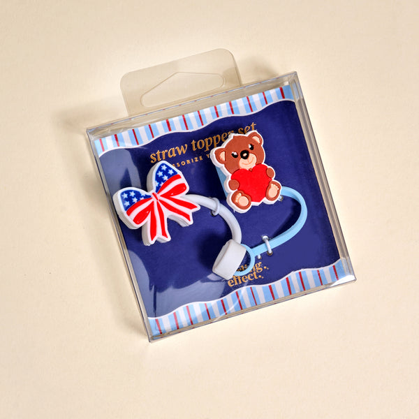 The Darling Effect’s Star Spangled Bear Straw Topper Set includes a blue and red bow with stars and a teddy bear holding a red heart, both attached to a flexible blue straw—cute, functional drinkware accessories.
