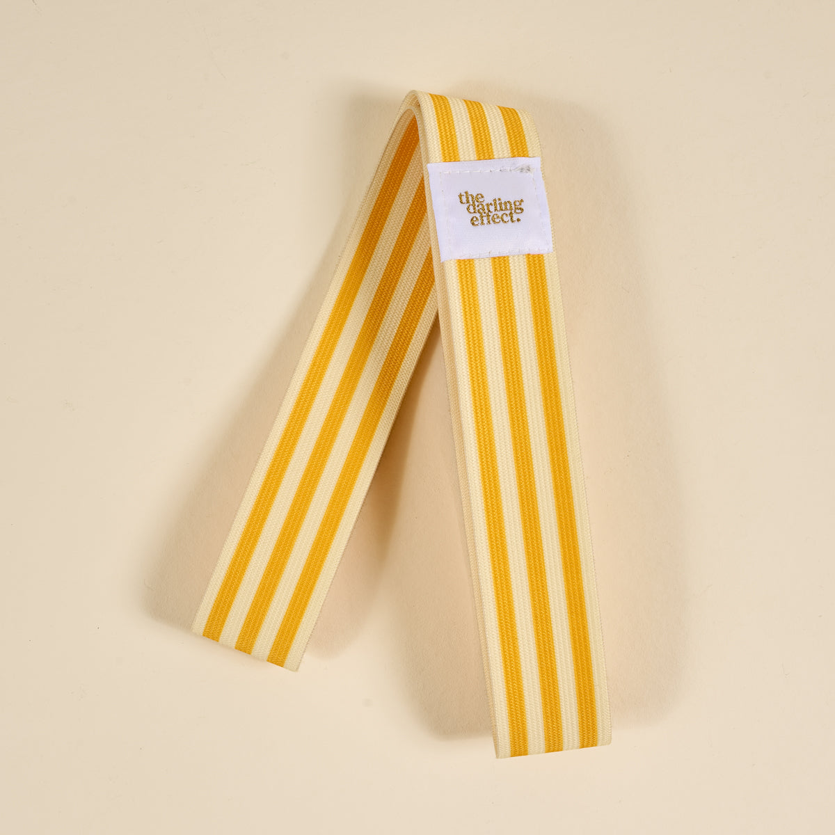 The Darling Effect Put Towel Band-Stripes Yellow Stay features yellow and cream stripes, a white tag with the logo, and works as a stylish, no-slip towel holder for your beach chair on sunny days.
