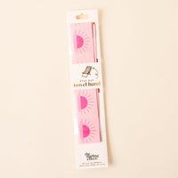 A packaged Put Towel Band-Sunshine Pink Stay by the-darling-effect, designed as a no-slip towel holder with a pink sun design; white packaging features gold and black text plus the brand name at the bottom. Perfect for beach chairs!.