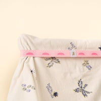 The-darling-effect's Put Towel Band-Sunshine Pink Stay secures a cream fabric with purple and yellow flowers. The pink straps have repeating sun motifs, all set against a plain beige background.