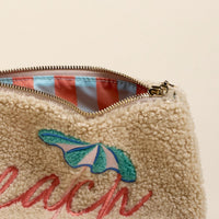 The Darling Effect's Beach Edit Teddy Pouch features cream sherpa fabric, a gold zipper, and “beach” in pink script beneath a blue and white embroidered beach umbrella. Inside, blue and pink striped lining completes this charming organizer.