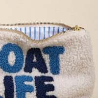 Close-up of the-darling-effect’s Boat Life Teddy Pouch in cream sherpa with blue “BOAT LIFE” embroidery. A partially open zipper reveals its cozy blue and white striped lining.