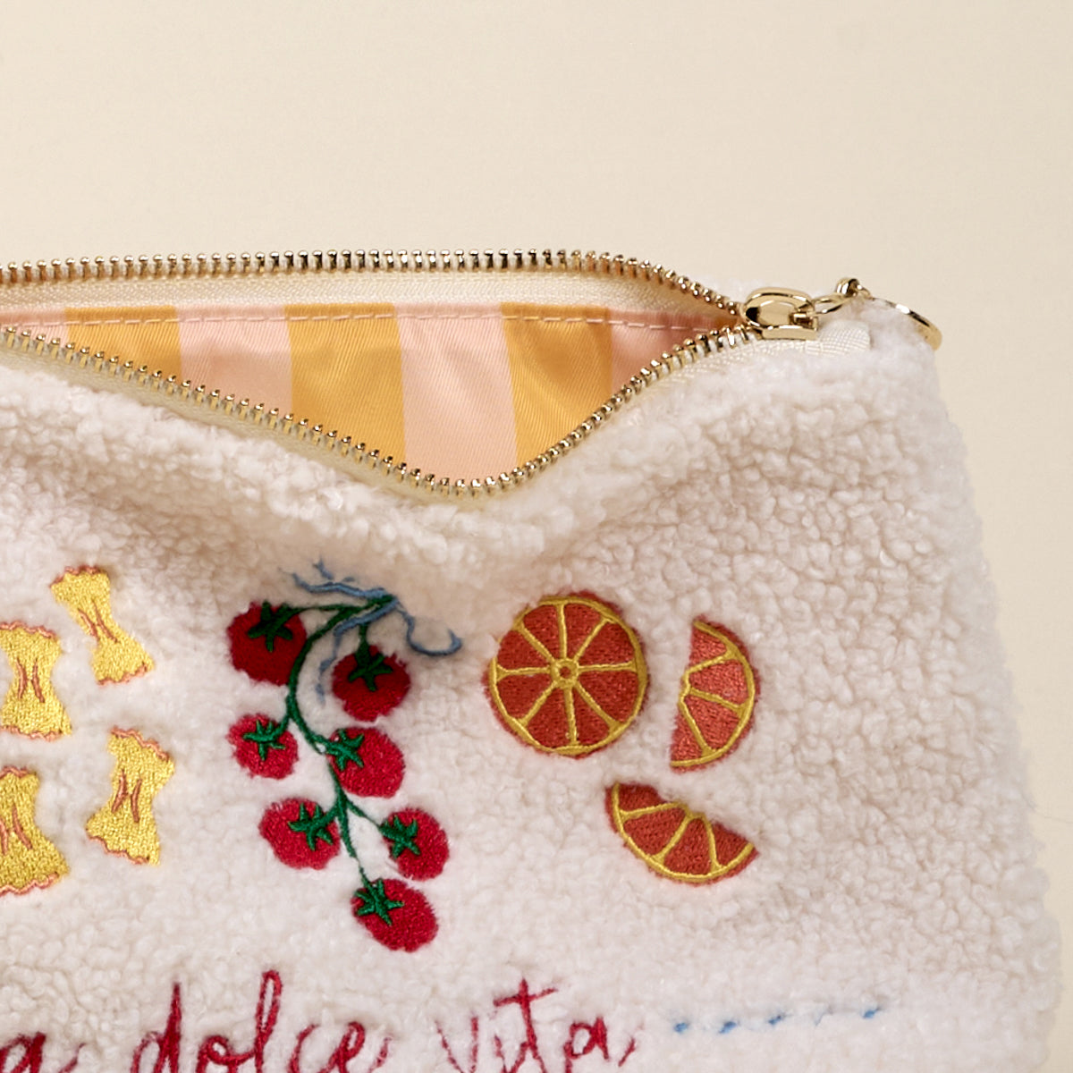 A close-up of The Darling Effect’s La Dolce Vita Teddy Pouch shows fuzzy white sherpa fabric embroidered with tomatoes, oranges, and pasta, and a partially open zipper revealing its pink and yellow striped lining.