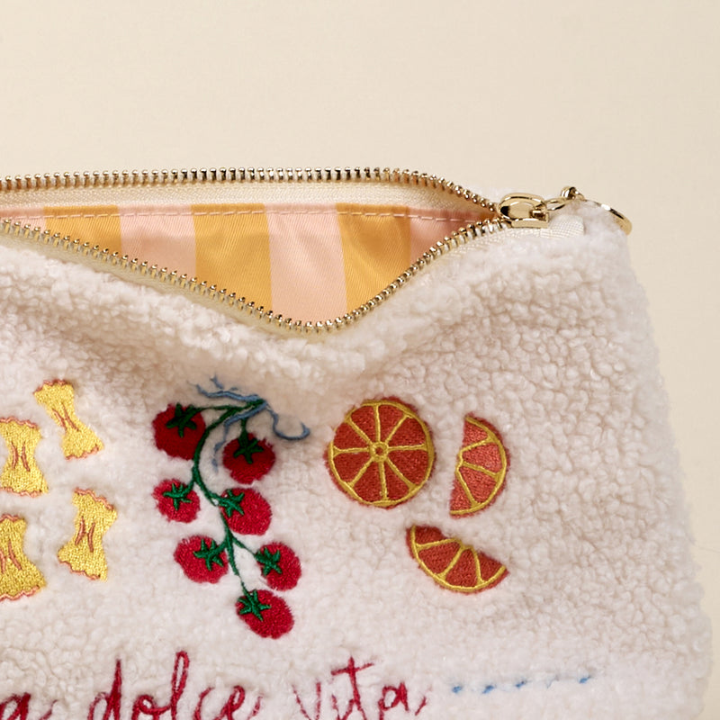 A close-up of The Darling Effect’s La Dolce Vita Teddy Pouch shows fuzzy white sherpa fabric embroidered with tomatoes, oranges, and pasta, and a partially open zipper revealing its pink and yellow striped lining.