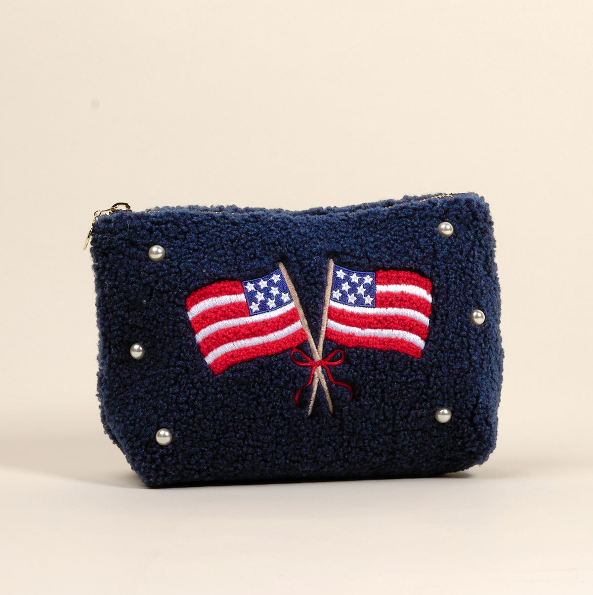 The Darling Effect’s American Flags Teddy Pouch is a blue textured sherpa zip pouch, accented with pearls and two crossed embroidered American flags tied with a red ribbon—an eye-catching accessory.