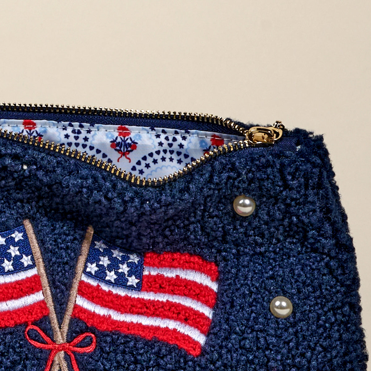 The Darling Effect’s American Flags Teddy Pouch is a navy textured sherpa pouch with embroidered flags, a red bow, pearl accents, and gold zipper, lined inside with white star and small red motif print fabric.