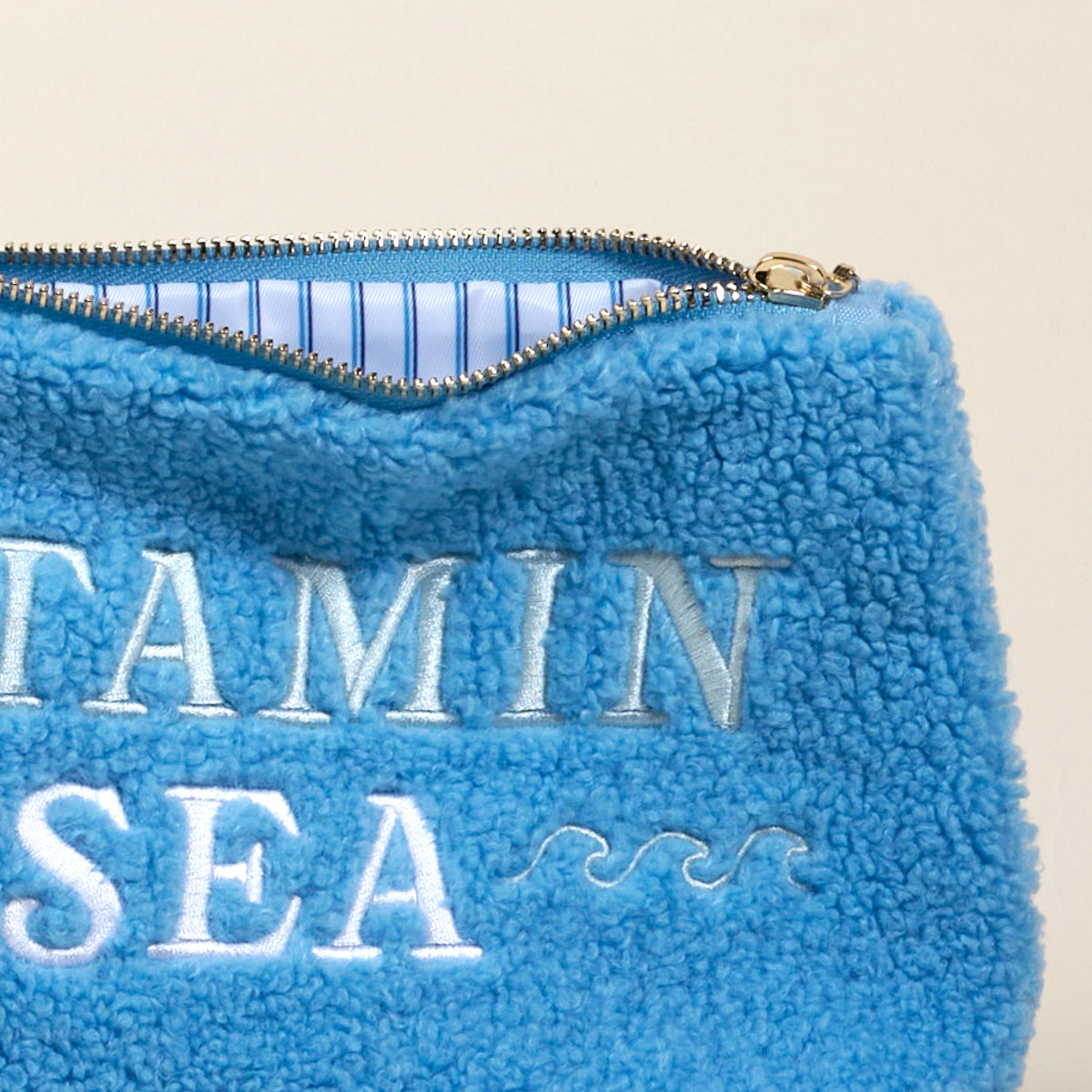 A close-up of The Darling Effect's Vitamin Sea Teddy Pouch: a blue sherpa pouch with "VITAMIN SEA" and wave embroidery, lined with blue and white stripes—an irresistibly stylish accessory.
