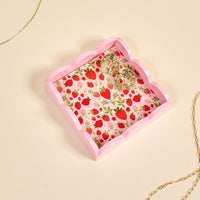 The Darling Effect's Strawberry Fields Scallop Trinket Tray features a pink scallop edge and strawberry print interior, shown holding gold hoop earrings and surrounded by gold chain necklaces on a cream background.