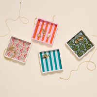 Four decorative accessory trays hold gold jewelry. Two have vertical stripes—pink and orange, or turquoise and white. The Darling Effect’s Strawberry Fields Scallop Trinket Tray shows a floral pattern, while another tray has a dark green design with blue flowers.