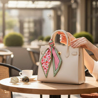 A person adjusts a white handbag with boutique charm, featuring The Darling Effect’s Strawberry Fields Twirl & Tie Scarf Accessory tied to its handle on a café table, next to sunglasses and a cup of coffee, in an outdoor sunny setting.