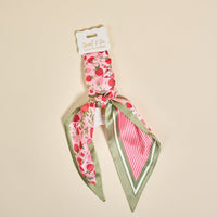 A square scarf from The Darling Effect, the Strawberry Fields Twirl & Tie Scarf Accessory features a charming strawberry print and green border. Folded neatly and attached to a branded cardboard tag, it’s displayed on a beige background. This boutique piece offers versatile styling options for any occasion.