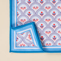 The Darling Effect’s All-American Darling Waffle Tea Towel features blue-edged trim and a patriotic pattern of heart-shaped blue stars, flags, and red flowers. One corner is folded to show the reverse side of this absorbent kitchen towel.