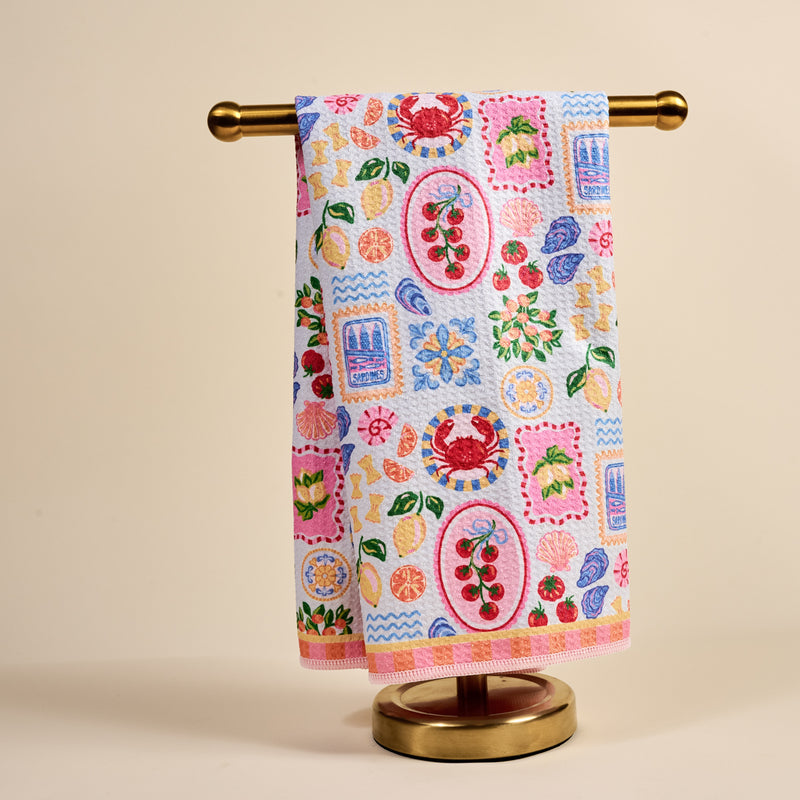 The Darling Effect's Ciao Bella Waffle Tea Towel, adorned with vibrant fruit and seafood prints, hangs neatly on a gold metal stand against a plain beige background.