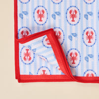 The Darling Effect Harbor House Waffle Tea Towel features a red border, blue stripes, and repeating red lobster and blue shell patterns. One corner is folded to display the underside of this absorbent kitchen towel.