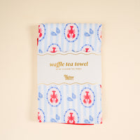A folded Harbor House Waffle Tea Towel by The Darling Effect features a blue and red lobster and seashell pattern, wrapped in a label, displayed on a light beige background.