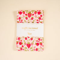 The Strawberry Fields Waffle Tea Towel by The Darling Effect features a vibrant strawberry design. Folded and wrapped in a white band labeled with the brand and product name, the absorbent towel rests on a solid light beige background.