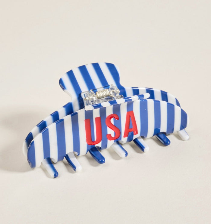 The USA Word Up Claw Clip by The Darling Effect is a cute hair accessory featuring blue and white stripes with bold red “USA” lettering on the front, making it stand out beautifully when placed on a light-colored surface.