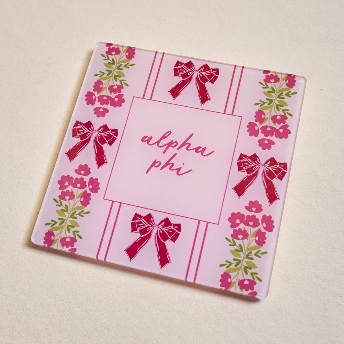 The Darling Effect’s Alpha Phi Acrylic Coaster features a pink striped background with “alpha phi” in cursive, accented by red bows and pink flowers with green leaves—an ideal sorority gift.