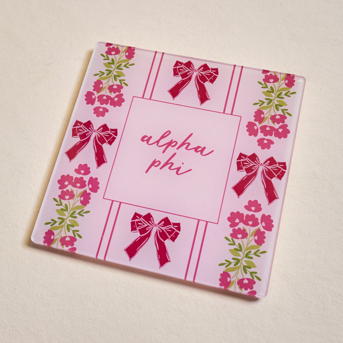 The Darling Effect’s Alpha Phi Acrylic Coaster features a pink striped background with “alpha phi” in cursive, accented by red bows and pink flowers with green leaves—an ideal sorority gift.