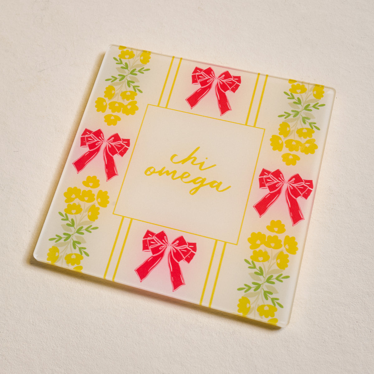 The Darling Effect’s Chi Omega Acrylic Coaster features a white square design with yellow floral patterns, red bows at the edges, and "chi omega" in yellow cursive—ideal as a sorority coaster or thoughtful gift.