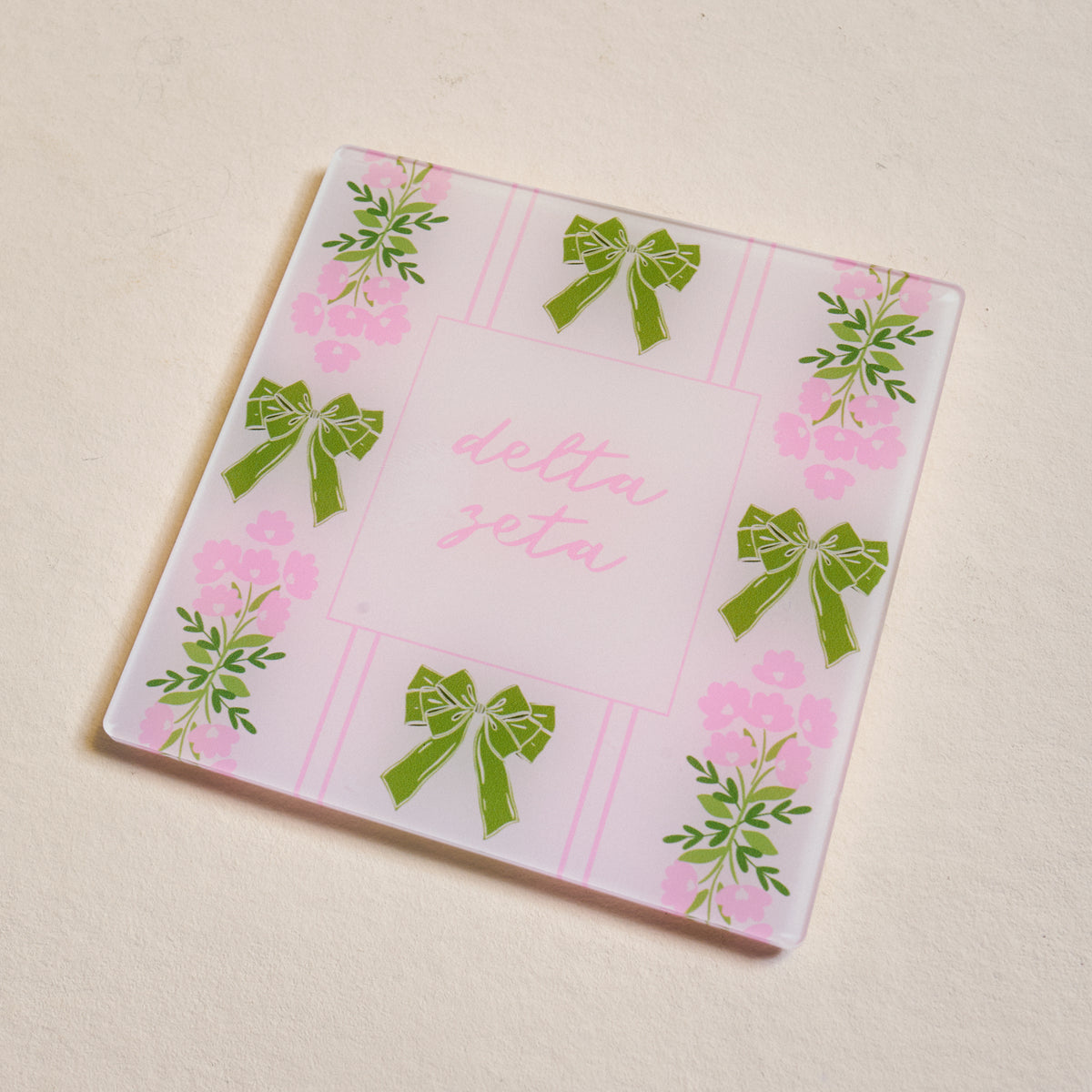 The Darling Effect’s Delta Zeta Acrylic Coaster features a light beige background, pink stripes, green bows, floral designs, and “Delta Zeta” in pink cursive—perfect for chapter pride or as a Big/Little gift.
